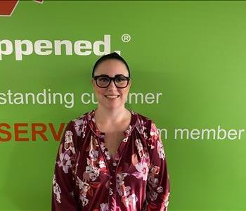 Female employee Sarah Raymond standing in front of a green wall with the SERVPRO logo and mission statement below it.